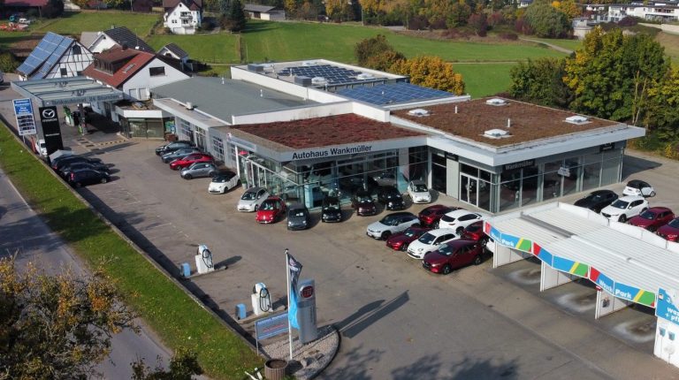 Das Autohaus mit mehreren Fahrzeugen und modernen Gebäuden, umgeben von Grünflächen. Bildwinkel entspricht einer Drohnenaufnahme.