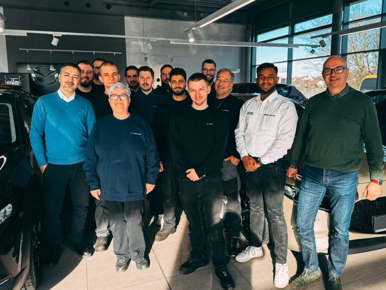 Eine Gruppe von 14 Personen in einem Autohaus, unterschiedliche Altersgruppen, lächelnd. Es stellt einen kleinen Auszugs des Teams dar.