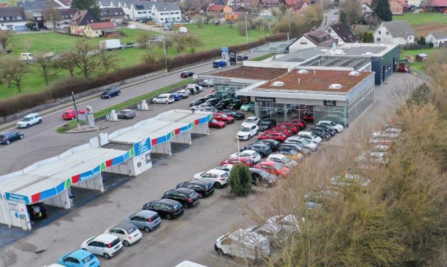 Parkplatz mit vielen Autos und einem Gebäuden inklusive der Waschanlage. Der Winkel entspricht einer Drohnenaufnahme.