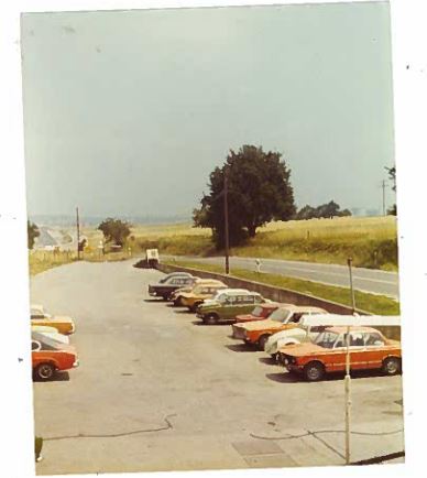 Parkplatz mit mehreren Autos, größtenteils orange, umgeben von Wiese und Straße. Es zeigt eine Aufnahme des Autohauses von früher.