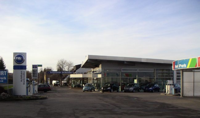 Autohaus mit modernen Glasfassaden und mehreren Fahrzeugen auf dem Gelände. Es beinhaltet das alte FIAT Logo.