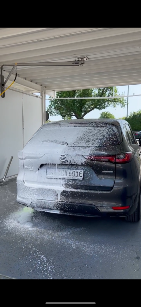 Schwarzes Auto, das mit Schaum in der Waschanlage besprüht wird.