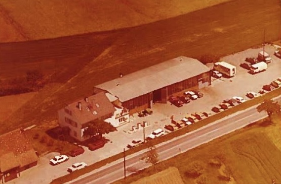 Luftaufnahme eines älteren Gebäudes mit vielen Fahrzeugen und einem Parkplatz. Es zeigt das Autohaus von früher in den Anfangszeiten.