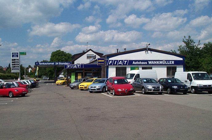 Autohaus mit altem FIAT Logo mit verschiedenen älteren Fahrzeugen auf dem Parkplatz und wolkigem Himmel.
